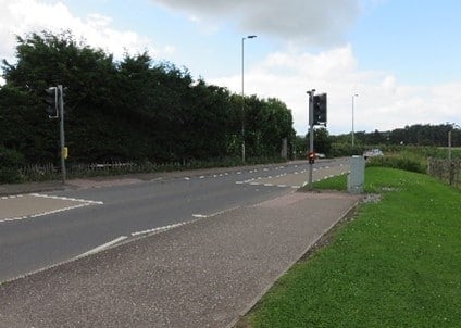 An image if a road with a pavement and a traffic lights crossing.