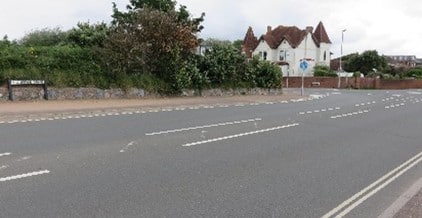 A road with a hedge and a house on the other side.