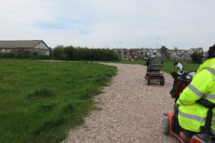 People using mobility scooters on a gravel path with grass on either side.