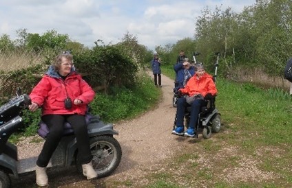 People using disability scooters on a path near to a hedge and trees.