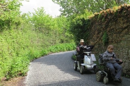 People using disability scooters on a road with high hedges on the sides.