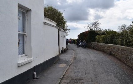 A road with a white house on the left and a low wall on the right.