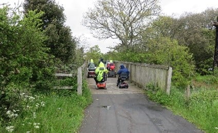People using disability scooters on a wooden boardwalk with trees on either side.